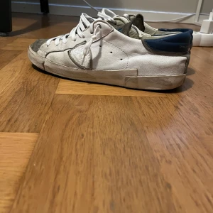 Vita sneakers från Philippe Model med blå detaljer - Säljer ett par vita Philippe Model sneakers med blå detaljer vid hälen och grå mocka vid tån. Skorna har snörning och en platt sula. Klassisk design med rund tå och märkets logga på sulan.