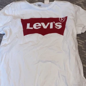 Vit t-shirt med röd Levi's-logga - Vit t-shirt från Levi's med den klassiska röda loggan tryckt på bröstet. Rund halsringning och korta ärmar. Perfekt för en avslappnad och stilren look.