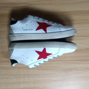 Vita sneakers med röd stjärna från Golden Goose - Säljer ett par vita sneakers från Golden Goose med en röd stjärna på sidan och svarta detaljer vid hälen. Skorna har snörning och en klassisk rund tå. Perfekta för dig som gillar en sportig och trendig stil.