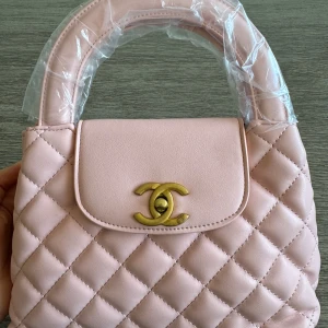 Ljusrosa quiltad handväska  - Säljer en ljusrosa quiltad handväska med guldfärgad logga framtill. Väskan har ett klassiskt rutmönster och ett handtag upptill. Perfekt accessoar för att lyfta din outfit. Helt ny!