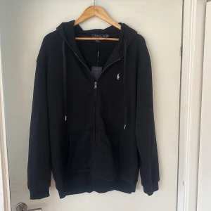 Svart hoodie med dragkedja från Polo Ralph Lauren - Svart hoodie från Polo Ralph Lauren med dragkedja framtill och klassisk logga på bröstet. Tröjan har huva med dragsko och två fickor framtill. Perfekt för en avslappnad stil.
