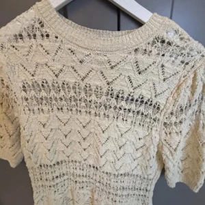 Beige stickad topp med spetsdetaljer - Säljer en beige stickad topp med korta ärmar och vackert spetsmönster. Toppen har rund halsringning och ett luftigt, virkat utseende som ger en bohemisk känsla. Perfekt att styla till jeans eller kjol.