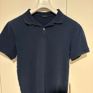 Mörkblå pikétröja från Massimo Dutti - Säljer en klassisk mörkblå pikétröja från Massimo Dutti med krage och knappslå. Perfekt för en stilren och avslappnad look. Tröjan har korta ärmar och är tillverkad i ett mjukt bomullsmaterial.