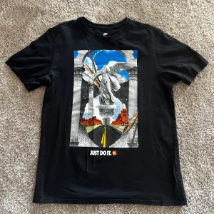 Svart t-shirt med tryck från Nike - Svart t-shirt från Nike med stort grafiskt tryck framtill som föreställer en staty, pelare och landskap. Texten 'Just Do It.' under motivet. Klassisk passform och rund halsringning.