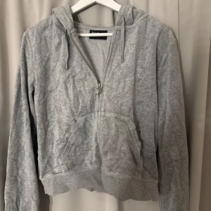 Grå hoodie med dragkedja från Juicy Couture - Säljer en klassisk grå hoodie från Juicy Couture med dragkedja framtill och huva med snören. Tröjan har långa ärmar, fickor på magen och en liten broderad logga på bröstet. Perfekt för en avslappnad stil.