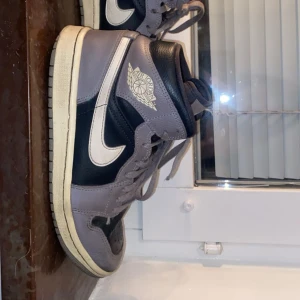 Nike Air Jordan 1 High i svart och grått - Säljer dessa för jag ej använder dem längre men dem är väldigt fina och perfekt året om. De är ett par klassiska Nike Air Jordan 1 High i svart och grått med vita detaljer och ikonisk vinge-logga på sidan. Skorna har snörning och en vit sula. Perfekta för dig som gillar streetwear och sneakers med retrostil.