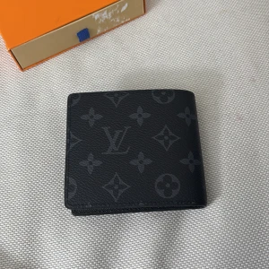 Svart plånbok från Louis Vuitton - Snygg svart plånbok från Louis Vuitton med klassiskt monogrammönster. Plånboken har flera kortfack, ett fönster för ID och är tillverkad i skinn. Perfekt för dig som vill ha en stilren accessoar med lyxig känsla.                 Pris 300kr. 