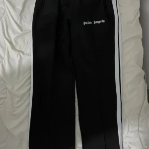 Svarta byxor med vita ränder från Palm Angels - Säljer ett par svarta byxor från Palm Angels med vita ränder längs sidorna och logga tryckt på vänster sida. Byxorna har raka ben och sidofickor, perfekt för en sportig och trendig look.
