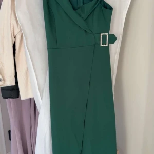 Grön långklänning från Trendyol med bälte - Säljer en elegant grön långklänning från Trendyol i storlek 36. Klänningen har en snygg omlottkrage och ett dekorativt bälte med glittrande spänne i midjan. Perfekt för dig som vill sticka ut med en stilren look.