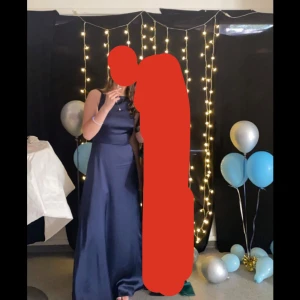 Mörkblå långklänning med spetsdetaljer på ryggen - Elegant mörkblå långklänning i satin med v-ringad rygg och spetsdetaljer. Klänningen är ärmlös och har en slits på sidan för extra stil. Perfekt för bal eller festliga tillfällen. Använd endast en gång och det var på min bal!