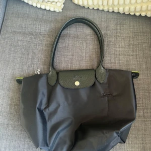 Svart handväska från Longchamp - Säljer en klassisk svart handväska från Longchamp med svarta handtag och lock i skinn. Väskan har en tryckknapp och dragkedja upptill. Perfekt storlek för vardagens alla prylar och enkel att bära över axeln. 