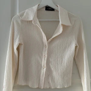 Beige skjorta med struktur från Cider. OBS! Den har ett läppstift märke vid armen. - Säljer en beige skjorta från Cider i storlek S. Skjortan har lång ärm, klassisk krage och knäppning framtill. Materialet är strukturerat och ger en snygg och modern look. Perfekt att styla med jeans eller kjol.