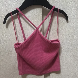 Rosa ribbad croppad linnetopp med tre smala axelband - Säljer en rosa croppad linnetopp med ribbad struktur och smala korsade axelband. Perfekt för varma dagar eller som ett lager under en skjorta. Enkel och stilren design som passar till mycket.