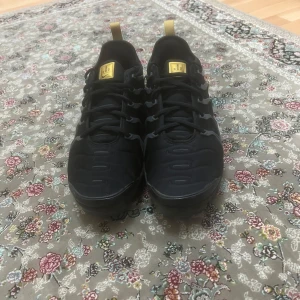 Svarta Nike Air Max Plus sneakers - Säljer ett par svarta Nike Air Max Plus sneakers med gulddetaljer och synlig Nike-logga på sidan. Skorna har snörning, platt sula och en sportig design med vågformade detaljer längs sidorna. Perfekta för dig som gillar streetwear och vill sticka ut.