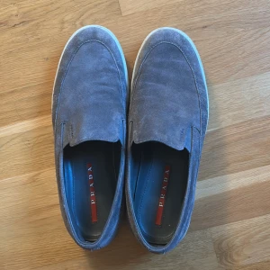 Grå mocka loafers från Prada - Säljer ett par stilrena grå loafers från Prada i mjuk mocka med vit platt sula. Skorna har diskret Prada-logga på sidan och röd detalj på innersulan. Perfekta för dig som gillar klassisk och clean stil. Såklart äkta! 
