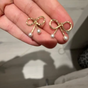 Guldiga rosettörhängen med pärlor - Säljer ett par söta örhängen i form av rosetter i guldton med små vita pärlor som hänger ned. Perfekta för att ge en gullig och elegant touch till din stil.