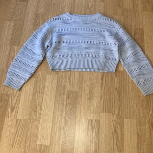 Ljusblå croppad stickad tröja från H&M - Säljer en ljusblå croppad stickad tröja från H&M. Tröjan har ett hålstickat mönster och långa ärmar. Perfekt för lager-på-lager och ger en trendig look. Ribbstickade muddar vid ärmslut och nederkant.