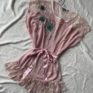 Ljusrosa klänning med spets och paljetter från Cotton Club - Supersöt ljusrosa klänning med spetsdetaljer upptill och nertill, samt glittriga paljetter och pärlor framtill. Satinband i midjan som knyts och volangkant nedtill. Perfekt för dig som gillar romantisk och feminin stil. Bältet har en stor svart fläck men går att byta ut(sista bilden)