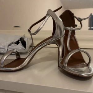 Silverfärgade sandaler med klack från H&M - Säljer ett par stilrena sandaler med klack från H&M i metallic silver. Skorna har öppen tå, smala remmar och spänne runt vristen. Perfekta för dig som vill sticka ut med en glansig look.