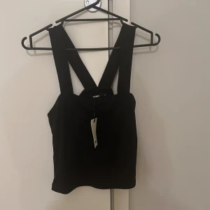 Svart croppad korsettopp - Säljer en svart croppad topp med breda axelband som korsas i ryggen. Toppen har en stilren och modern design, perfekt att styla till olika outfits. Materialet känns mjukt och stretchigt.