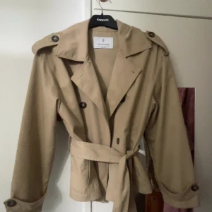 Beige trenchcoat från Stradivarius - Säljer en beige trenchcoat från Stradivarius med klassisk krage, knappar och bälte i midjan. Jackan har axelklaffar och långa ärmar med knappar vid ärmslut. Perfekt för en stilren look under övergångssäsonger. Fått en liten fläck därav priset 
