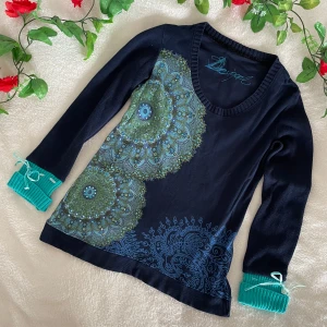 Mörkblå mönstrad tröja från Desigual - Säljer en unik mörkblå tröja från Desigual med grönt och blått mandalamönster framtill. Tröjan har långa ärmar med turkosa ribbade muddar och dekorativa band. Perfekt för dig som gillar färg och detaljer!