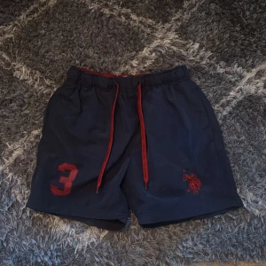 Mörkblå badshorts från U.S. Polo Assn. - Mörkblå badshorts från U.S. Polo Assn. med röd snörning i midjan, röd siffra 3 på ena benet och broderad logga. Shortsen har elastisk midja och bakficka. Perfekta för en avslappnad stil.