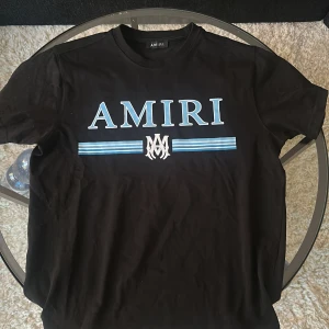 Svart t-shirt från Amiri med tryck - Svart t-shirt från Amiri med stort blått och vitt logotryck på bröstet. Klassisk rund halsringning och normal passform. Tillverkad i mjuk bomull för en skön känsla.