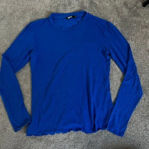 Blå långärmad topp från Only - En enkel och stilren blå långärmad topp från Only. Modellen har rund halsringning och vågig kant nertill och vid ärmslut. Perfekt att styla till jeans eller kjol för en clean look.