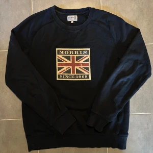 Morris Tröja Navy M - Slitningarna som förekommer på bilden är enligt design. Svart sweatshirt från Morris med stort Union Jack-tryck och texten 'Morris Since 1968' på bröstet. Klassisk rund halsringning och ribbade muddar vid ärmslut och nederkant. Perfekt för en stilren och avslappnad look.