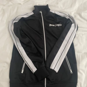 Svart track jacket från Palm Angels - Säljer en svart track jacket från Palm Angels med vita ränder längs ärmarna och dragkedja framtill. Jackan har hög krage, ribbade muddar och två fickor med dragkedja. Klassisk logga på bröstet. Storlek XS men passar som S/M, priset kan diskuteras