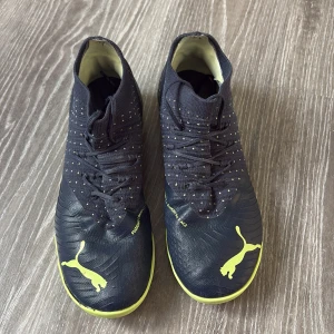 Svarta och neongula fotbollsskor från Puma - Säljer ett par svarta fotbollsskor från Puma med neongula detaljer och logga. Skorna har snörning och en låg profil med mönstrad sula för bra grepp på inomhusplan. Perfekta för dig som vill sticka ut på planen!