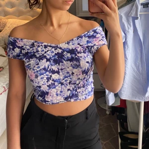 Blommig croppad topp i blått och lila - Säljer en croppad topp med blommigt mönster i blått, lila och vitt. Toppen kan du både ha som offshoulder och med vanliga axlar. 