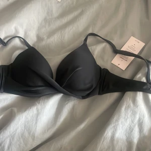 Svart bikini överdel från Nelly - Säljer en svart push up bikini-bh från Nelly med twistad front och justerbara axelband. Perfekt för stranden eller poolen, med snygg och enkel design. Stängs med spänne i ryggen. Använd en gång. Ordinarie pris 250 kr. 