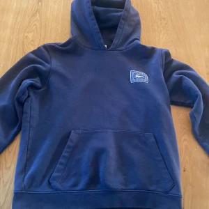 Blå hoodie från Lacoste - Säljer en blå hoodie från Lacoste med klassisk krokodillogga på ärmen och stort tryck på ryggen. Tröjan har magficka och huva, perfekt för en avslappnad stil. Mjuk och skön bomullskänsla.