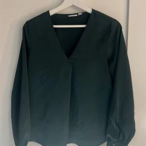 Mörkgrön satin blus - Stilren svart blus med djup v-ringning och långa ärmar. Ärmsluten har knappdetalj för en extra touch. Perfekt för dig som gillar en enkel men elegant look.