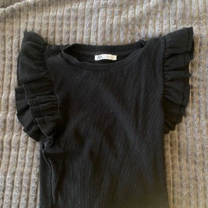 Svart ribbad topp med volangärmar från Zara - Säljer en svart ribbad topp från Zara med stora volangärmar som ger en cool och trendig look. Toppen är tight och har en rund halsringning. Perfekt för dig som vill sticka ut lite extra!