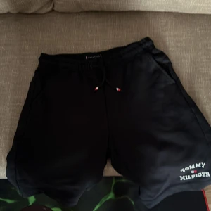 Svarta shorts från Tommy Hilfiger - Svarta shorts från Tommy Hilfiger med snörning i midjan och logga tryckt på ena benet. Mjukt material och fickor på sidorna samt en bakficka med liten flaggdetalj. Perfekta för avslappnade dagar.