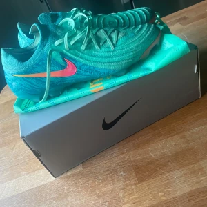 Nike Phantom GX II Elite LV8 FG fotbollsskor - Säljer ett par Nike Phantom GX II Elite LV8 FG i färgen Green Glow med rosa och orange swoosh. Skorna har snörning och en modern, strömlinjeformad design med rund tå och platt sula. Perfekta för spel på gräsplan. Har inte används då jag har varit skadad nyskick! Nypris ca 3200