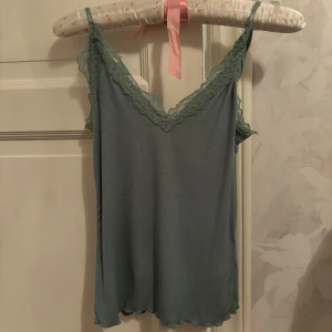 Grön linnetopp med spetsdetaljer - Säljer en grön linnetopp med smala axelband och vacker spets längs v-ringningen och ryggen. Toppen har en mjuk, ribbad struktur och är perfekt för varma dagar eller som lager under en kofta.