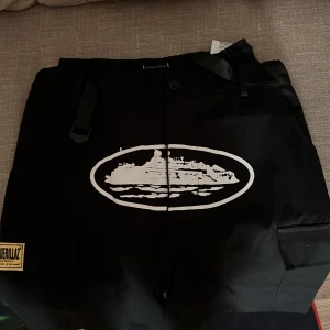 Svarta cargopants från corteiz  - Svarta cargopants med stort vitt tryck av ett fartyg på framsidan och flera fickor med lock. Byxorna har patchdetaljer och justerbart band i midjan. Perfekta för en streetwear-look.