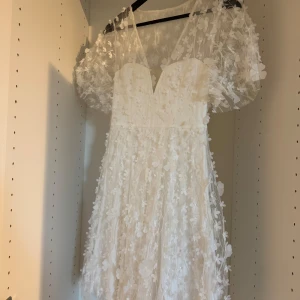 Perfekta studentklänningen  - Drömmig vit långklänning med v-ringning och puffiga korta ärmar. Klänningen är täckt av 3D-blommor som ger en romantisk och unik look. Perfekt för speciella tillfällen eller som studentklänning. Den är ifrån Bubbleroom