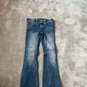Blå bootcut jeans från H&M - Säljer ett par klassiska blå bootcut jeans från H&M med låg midja och justerbar midja. Jeansen har fickor med lock och knapp bak samt snygga slitningar framtill. Perfekta för dig som gillar en retro vibe!