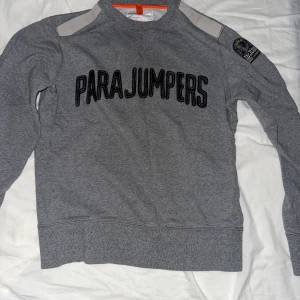 Grå sweatshirt från Parajumpers - Säljer en grå sweatshirt från Parajumpers med stor svart logga broderad över bröstet. Tröjan har rund halsringning och långa ärmar. Klassisk och stilren design med patch på ena ärmen.