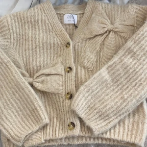 Beige kofta med rosett från Zara - Superfin oanvänd beige kofta från Zara i stickat material med prislappen kvar. Koftan har v-ringning, knappar framtill och en stor dekorativ rosett på båda sidorna. Perfekt för kyliga sommarkvällar💗💗
