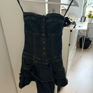 Mörkblå jeansklänning med volang och knappar - Säljer en mörkblå jeansklänning med  volang nertill, står xxs men passar xs. Klänningen har dekorativa knappar framtill och markerade sömmar som ger en snygg form. Säljer då den inte passade💕
