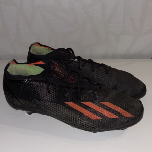 Svarta fotbollsskor från Adidas med röda ränder - Säljer ett par svarta fotbollsskor från Adidas med röda klassiska ränder på sidorna. Skorna har snörning och är tillverkade i syntetmaterial med låg profil och dobbar för bra grepp på planen.