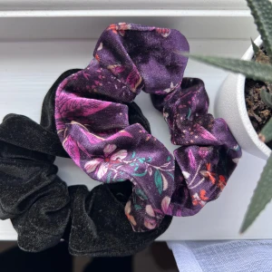 Två scrunchies i sammet och mönstrat tyg - Säljer två scrunchies, en i svart sammetsmaterial och en i lila sammetstyg med blommigt mönster i rosa, grönt och orange. Perfekta som accessoar för att piffa upp din frisyr. Aldrig använda.