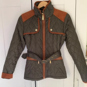 Quiltad jacka med bälte från Vince Camuto - Snygg quiltad jacka i olivgrönt med bruna detaljer och axelpartier. Jackan har lång ärm, dragkedja, knappar, bälte i midjan och dekorativa detaljer vid fickor och axlar. Perfekt för höst och vår. Köpte den i USA och har aldrig använt den. 