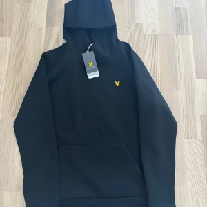 Svart hoodie från Lyle & Scott - Säljer en svart hoodie från Lyle & Scott med klassisk gul logga på bröstet. Tröjan har huva med dragsko och en stor magficka framtill. Perfekt för en avslappnad stil. Helt ny
